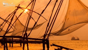 Traditional Chinese fishing nets silhouetted against an orange sunset on the Kochi waterfront, with a boat visible in the distance and the Brio Travels logo on the image. Kerala tour packages for family
