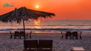 Sunset beach scene with two wooden lounge chairs under a large thatched palm-leaf umbrella, surrounded by tables and chairs on soft sand. The calm ocean reflects the warm orange glow of the setting sun, with a small boat visible in the distance. The 'Brio Travels' logo marks the tranquil, vacation-themed composition. Especially for Honeymoon in India
